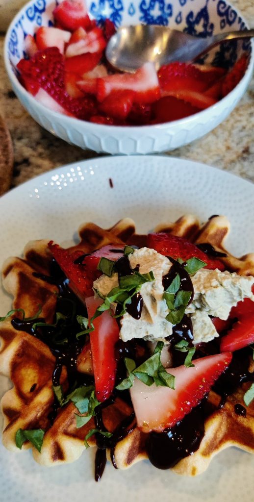 Waffles with chocolate sauce drizzled  on top, covered in sliced strateberries, a basil whipped cream and chopped fresh basil. 