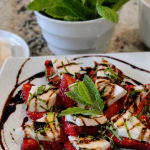 Strawberry caprese on a plate, layered slices of strawberries and mozarella topped with chopped mint and balsalmic glaze.