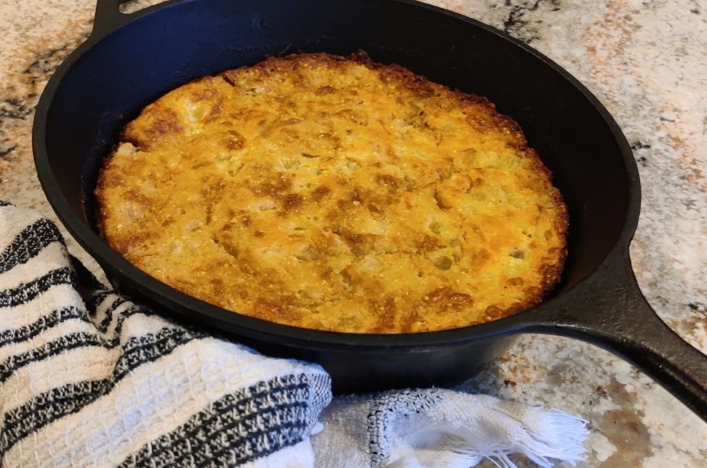 Golden green chile and cheese cornbread in a cast iron skillet on a countertop with a dish towel, showcasing its crispy edges.