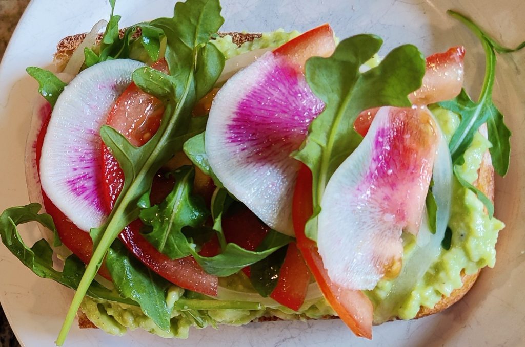 Sliced sourdough bread topped with mashed avocado, sliced tomatoes, colorful pink watermelon radishes and arugula.