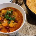 Vegan Portuguese bean soup topped with chopped parsley, served in a white bowl on the counter next to a cast iron skillet of cornbread.